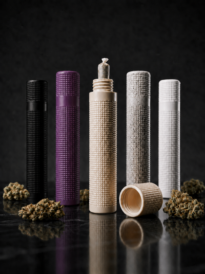 Fünf geruchshemmende Premium Joint Tubes von Strainlab in den Farben Schwarz, Lila, Beige, Grau gesprenkelt und Weiß. Die Hüllen stehen auf einer dunklen Oberfläche umgeben von Cannabis-Blüten. In der geöffneten, beigen Tube in der Mitte steckt ein fertig gerollter Joint.